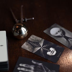 May include: A deck of tarot cards featuring black and white imagery, including a card labeled The Star with a starfish design and a card depicting a hand. A metallic spherical object with a pointed top sits next to the cards on a dark surface.