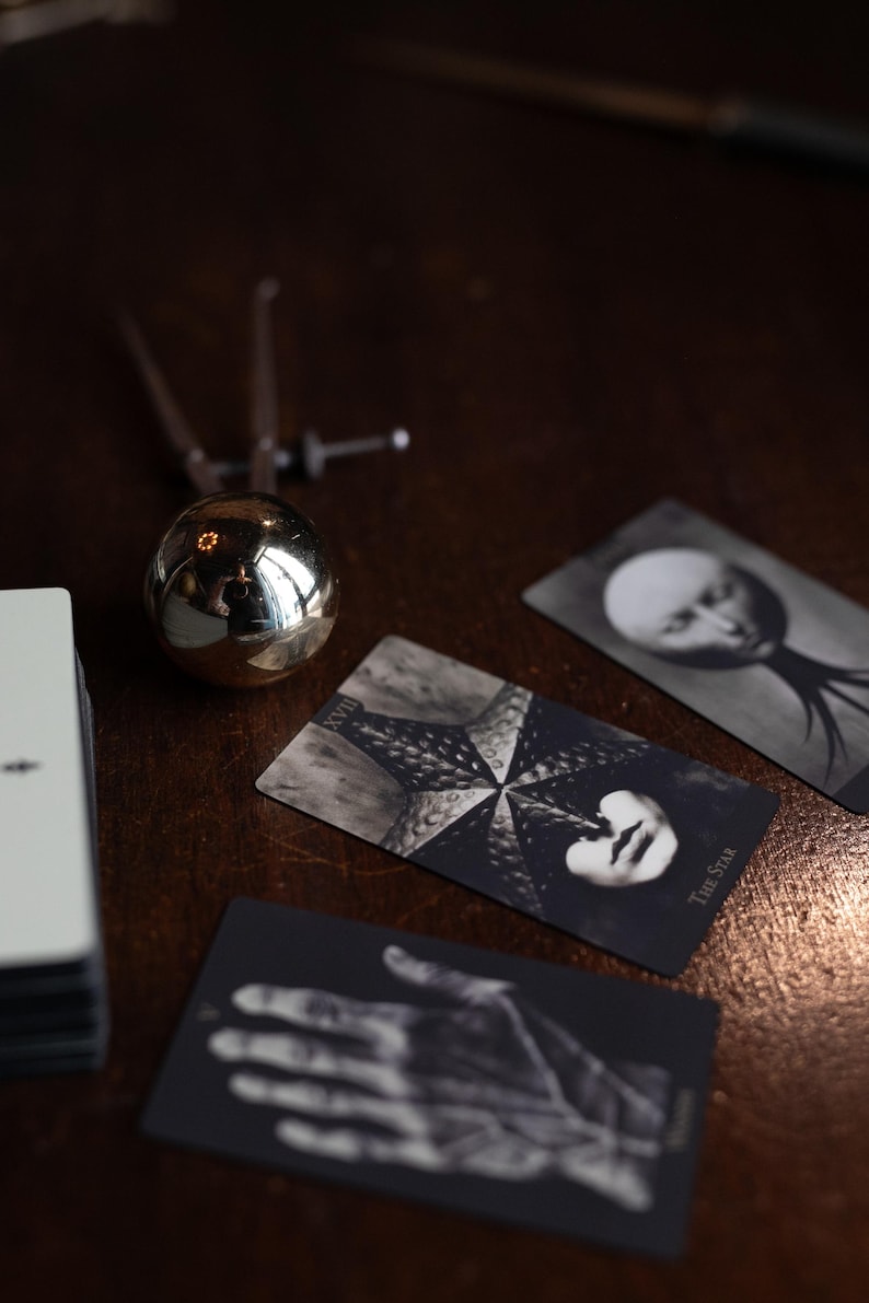 May include: A deck of tarot cards featuring black and white imagery, including a card labeled The Star with a starfish design and a card depicting a hand. A metallic spherical object with a pointed top sits next to the cards on a dark surface.