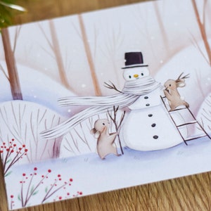 May include: A watercolor illustration of a snowman with a white scarf and a black top hat. Two small brown animals are helping to build the snowman. One animal is holding a stick and the other is standing on a ladder. The background is a snowy forest with brown trees and red berries.