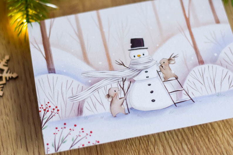 May include: A watercolor illustration of a snowman with a white scarf and a black top hat. Two small brown animals are helping to build the snowman. One animal is holding a stick and the other is standing on a ladder. The background is a snowy forest with brown trees and red berries.