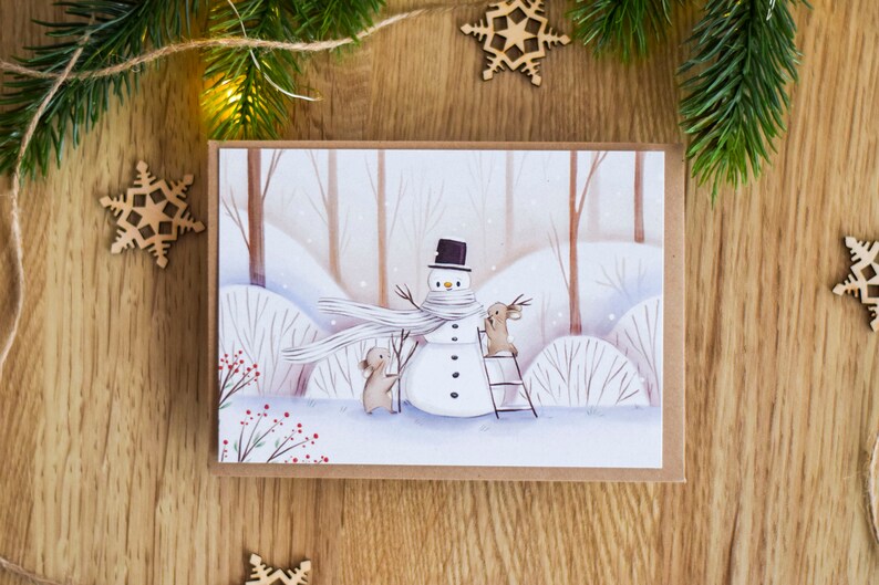 May include: A winter scene card with a snowman wearing a white scarf and a black hat. Two brown rabbits are building the snowman. One rabbit is on a ladder and the other is holding a stick. The background is a snowy forest with brown trees and white snow.