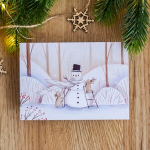 May include: A watercolor illustration of a snowman with a black top hat and a white scarf. Two brown rabbits are building the snowman. One rabbit is holding a branch and the other is standing on a ladder. The illustration is on a white card with a woodgrain background.