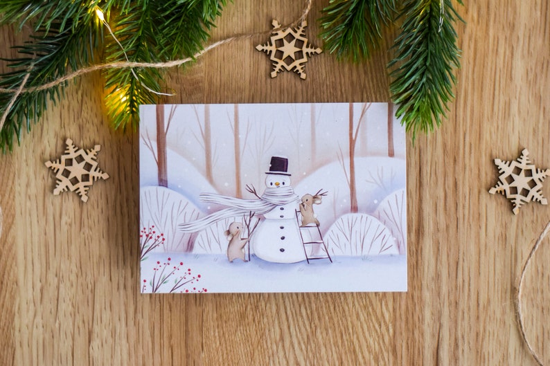 May include: A watercolor illustration of a snowman with a black top hat and a white scarf. Two brown rabbits are building the snowman. One rabbit is holding a branch and the other is standing on a ladder. The illustration is on a white card with a woodgrain background.