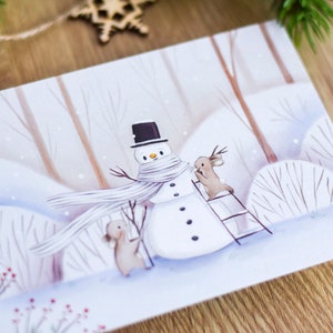May include: A winter scene with a snowman wearing a white scarf and a black top hat. Two brown rabbits are standing near the snowman, one is on a ladder. The background is a snowy forest with white and brown trees.