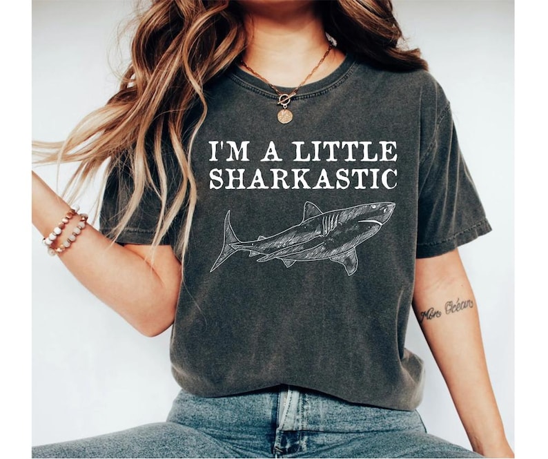 Peut inclure: Un t-shirt gris fonc&eacute; avec un graphique blanc d'un requin et le texte "I'm a little sharkastic".