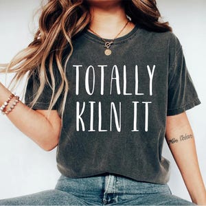 May include: A dark gray t-shirt with the text "TOTALLY KILN IT" printed in white.