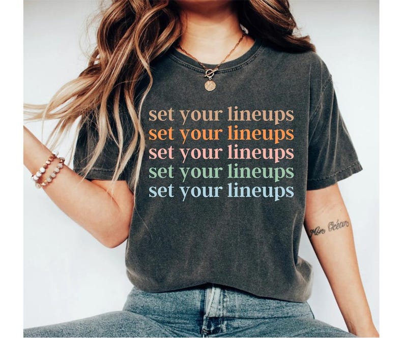 May include: A dark grey t-shirt with the text "set your lineups" repeated five times in different pastel colours. The text is centred on the shirt.