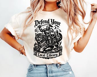 May include: Cream-colored t-shirt featuring a black and white graphic. The graphic depicts a knight riding a cat, with the text "Defend Your Local Library". The design includes a shield, sword, and a castle.
