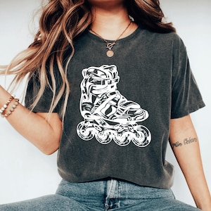 May include: Dark gray t-shirt featuring a white graphic of a roller skate. The graphic is a detailed illustration of the skate's components, including the boot, straps, and wheels. The shirt is a casual style.