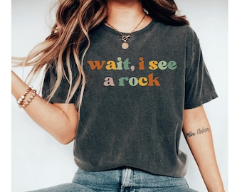 Op de afbeelding: Een antracietgrijs T-shirt met korte mouwen en de tekst "wait, i see a rock" gedrukt in een meerkleurig, retro-geïnspireerd lettertype. Het shirt heeft een ronde hals en een relaxte pasvorm, met woorden in oranje, groene en roze tinten.