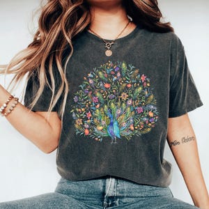 May include: Dark gray t-shirt featuring a vibrant peacock design. The peacock is depicted with a colorful array of feathers, flowers, and foliage. The shirt has short sleeves and a relaxed fit, suitable for casual wear.