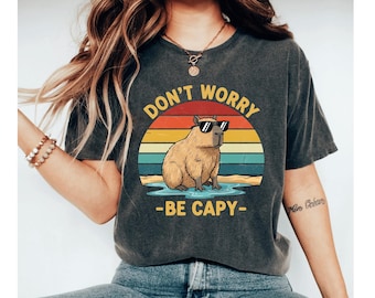 May include: Dark gray t-shirt featuring a capybara wearing sunglasses, set against a colorful sunset backdrop. The text "DON'T WORRY - BE CAPY" is printed above and below the capybara. A fun, casual design.
