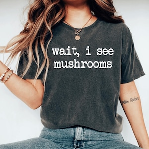 May include: Dark gray t-shirt with the text "wait, i see mushrooms" in white. The shirt is a crew neck style and appears to be made of a soft, comfortable material. The model is wearing blue jeans.