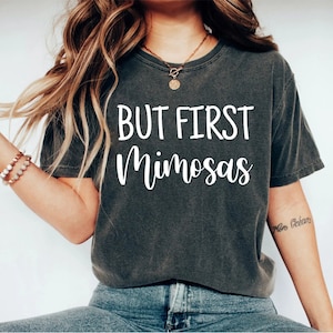 May include: A dark gray t-shirt with the text "BUT FIRST Mimosas" printed in white on the front.