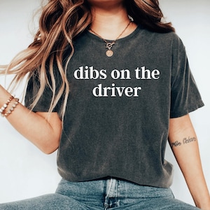 May include: Dark gray t-shirt with the text "dibs on the driver" in white. The shirt is a short-sleeved, crew neck style. The model is wearing blue jeans and a gold necklace.
