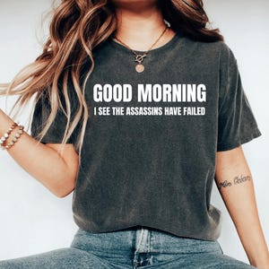 May include: Dark gray t-shirt with the text "GOOD MORNING I SEE THE ASSASSINS HAVE FAILED" in white. The shirt is a casual style, suitable for everyday wear.