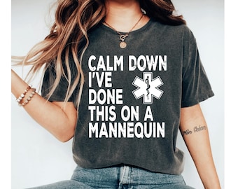 Może przedstawiać: Ciemnoszara koszulka z białym napisem "CALM DOWN I'VE DONE THIS ON A MANNEQUIN" i białym symbolem medycznym. Koszulka ma okrągły dekolt i krótkie rękawy.