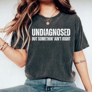 May include: Dark gray t-shirt with the text "UNDIAGNOSED BUT SOMETHIN' AIN'T RIGHT" in white block letters. The shirt is a crew neck style and appears to be made of a soft, comfortable material. The model is wearing blue jeans.