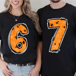 May include: Black t-shirts with large orange numbers 6 and 7. The numbers are filled with a spiderweb pattern and small black spiders. The numbers have a white outline and a stitched border, perfect for Halloween.