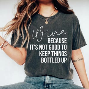 May include: A dark gray t-shirt with the text "Wine because it's not good to keep things bottled up" printed in white.