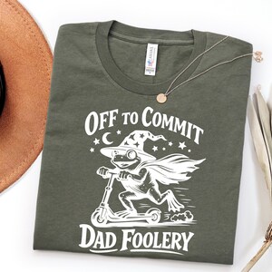 May include: Olive green t-shirt with the text "OFF TO COMMIT DAD FOOLERY". The graphic features a frog wearing a wizard hat and cape, riding a scooter. A brown hat and necklace are also in the image.