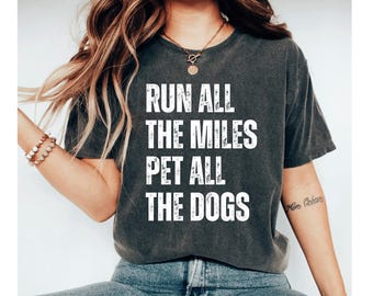 May include: Dark gray t-shirt with the white text "RUN ALL THE MILES PET ALL THE DOGS." The shirt is a crew neck and has short sleeves. The model is wearing a bracelet and necklace.