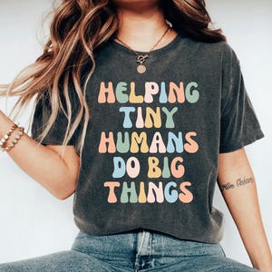 May include: Dark gray t-shirt with the text "HELPING TINY HUMANS DO BIG THINGS" in colorful, retro-style lettering. The shirt has a vintage, worn look.
