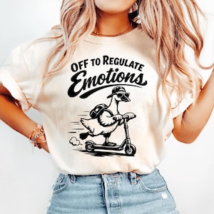 May include: Cream-colored t-shirt with a black graphic of a goose riding a scooter, wearing sunglasses and a hat. The text "OFF TO REGULATE Emotions" is above the goose. The shirt is paired with blue jeans.
