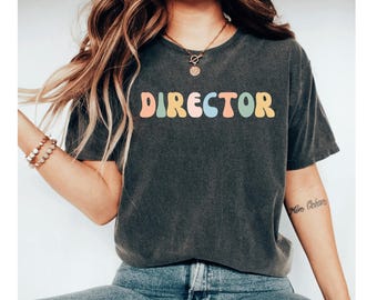 Puede incluir: Camiseta gris oscuro con la palabra "DIRECTOR" en letras coloridas de estilo retro. La camiseta tiene un aspecto vintage y está hecha de un material suave. El diseño es simple y llamativo.