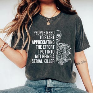 May include: A dark gray t-shirt with a white graphic of a skeleton with flowers. The text on the shirt reads "People need to start appreciating the effort I put into not being a serial killer."
