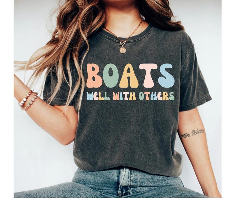 May include: A dark grey t-shirt with the text "BOATS WELL WITH OTHERS" in colourful, retro-style lettering.