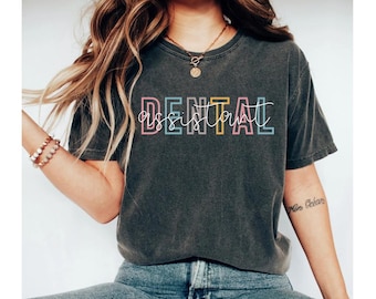 Op de afbeelding: Een donkergrijs T-shirt met de tekst "DENTAL ASSISTANT" in kleurrijke omrande letters.