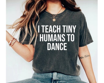 May include: A dark grey t-shirt with the text "I teach tiny humans to dance" printed in white on the front. The shirt is being worn by a person with long brown hair and a necklace.