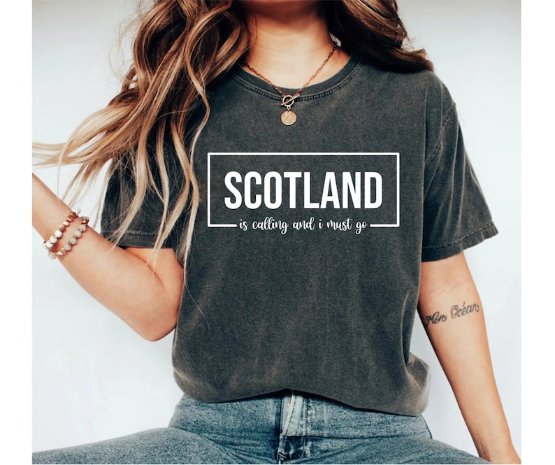 Pu&ograve; includere: Una maglietta grigia scura con testo bianco che dice "SCOTLAND - is calling and i must go".