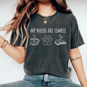 May include: A charcoal gray short-sleeve t-shirt featuring the white text MY NEEDS ARE SIMPLE above three line-art icons: a coffee cup with a heart, a paw print with a heart, and an open book with three small hearts floating above the pages.