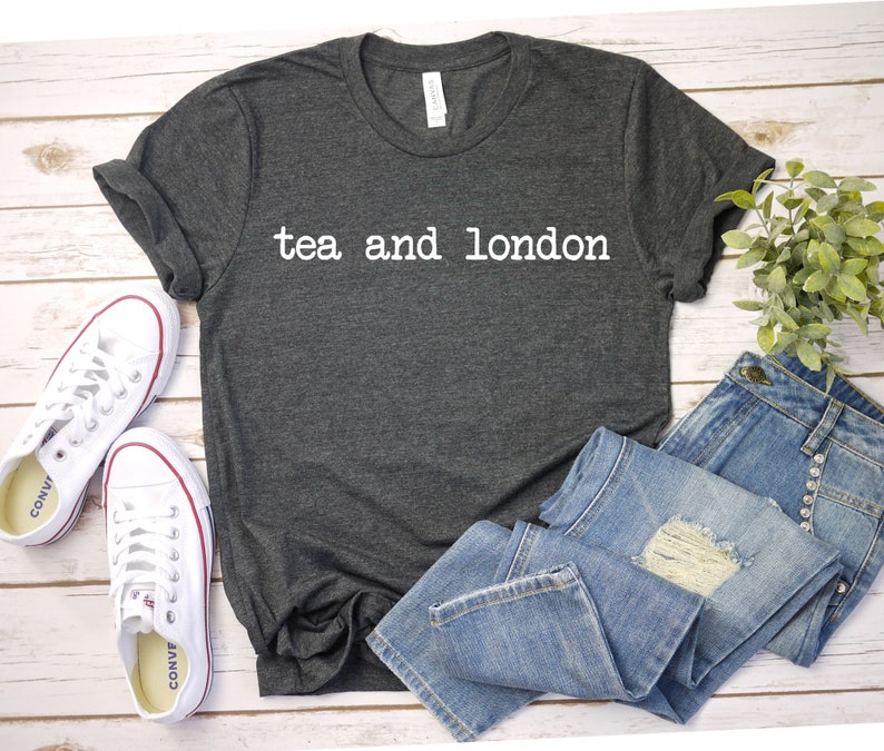 May include: A dark gray t-shirt with the text "tea and london" printed in white on the front. The shirt is laid flat on a white wooden surface with a pair of white sneakers and a pair of blue jeans.