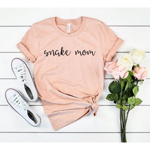 May include: A light pink t-shirt with the text "snake mom" printed in black on the front. The shirt is folded and laid flat on a white wooden surface. There are white sneakers and a bouquet of white and pink roses in the image.