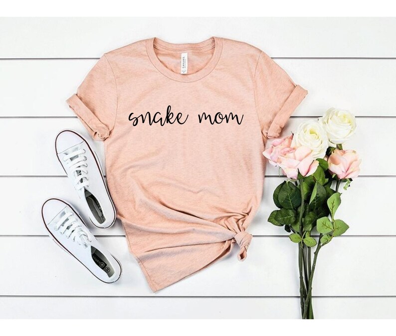 May include: A light pink t-shirt with the text "snake mom" printed in black on the front. The shirt is folded and laid flat on a white wooden surface. There are white sneakers and a bouquet of white and pink roses in the image.
