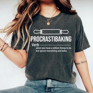 May include: A dark gray t-shirt with a white graphic design that says "PROCRASTIBAKING" and defines it as a verb: "when you have a million things to do but ignore everything and bake."