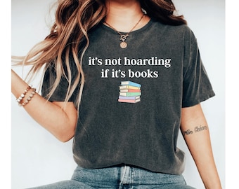 Puede incluir: Camiseta gris oscuro con la frase "it's not hoarding if it's books" sobre una pila de libros coloridos. Los libros son de color azul, verde, amarillo, naranja y morado. La camiseta es de estilo casual.