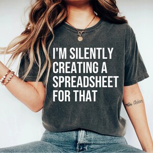 May include: Dark gray t-shirt with the text "I'M SILENTLY CREATING A SPREADSHEET FOR THAT" in white block letters. The shirt has short sleeves and a crew neck.