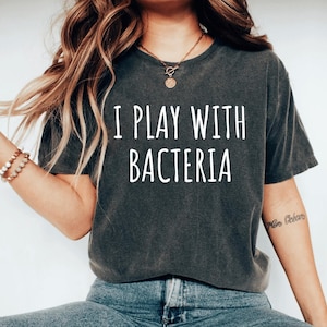 May include: A dark gray t-shirt with the text "I Play With Bacteria" printed in white on the front. The shirt is being worn by a person with long brown hair.
