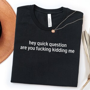 May include: Black t-shirt with the text "hey quick question are you fucking kidding me" in white. A gold necklace with a pendant is draped across the shirt. A brown hat is visible in the upper left corner.