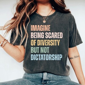 May include: Dark gray t-shirt with the text "IMAGINE BEING SCARED OF DIVERSITY BUT NOT DICTATORSHIP" in colorful block letters. The shirt is a casual style, suitable for everyday wear.