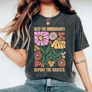 May include: Dark gray t-shirt with a floral design and the text "KEEP THE IMMIGRANTS DEPORT THE RACISTS". The graphic features pink, yellow, orange, and white flowers with green leaves. The shirt has a vintage, worn look.