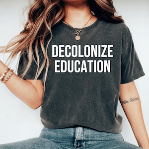 May include: Dark gray t-shirt with the words "DECOLONIZE EDUCATION" in bold white letters. The shirt has a vintage, worn look. The model is wearing blue jeans and a gold necklace.