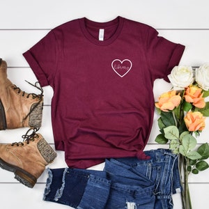 Puede incluir: Una camiseta de color granate con un coraz&oacute;n blanco y la palabra "library" dentro del coraz&oacute;n. La camiseta est&aacute; sobre una superficie blanca con un par de botas marrones, unos vaqueros azules y un ramo de flores.