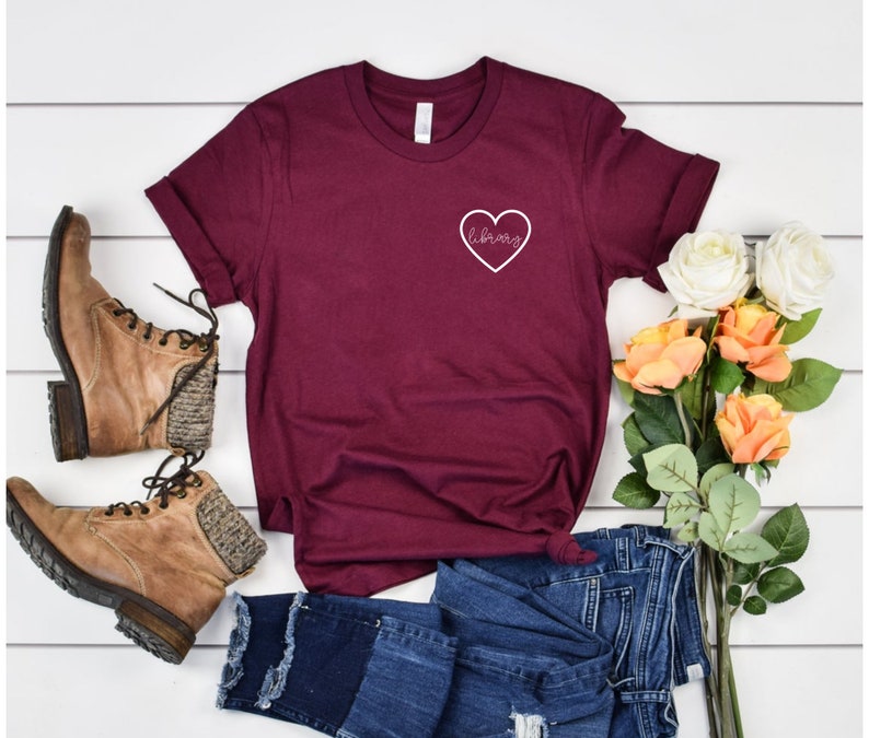 Puede incluir: Una camiseta de color granate con un coraz&oacute;n blanco y la palabra "library" dentro del coraz&oacute;n. La camiseta est&aacute; sobre una superficie blanca con un par de botas marrones, unos vaqueros azules y un ramo de flores.