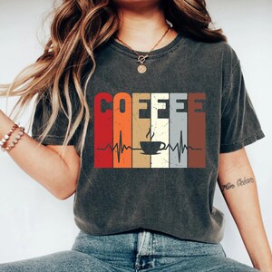 May include: A dark gray t-shirt with a vintage-style graphic of a coffee cup and the word "COFFEE" in a rainbow of colors. The graphic also includes a stylized heartbeat.