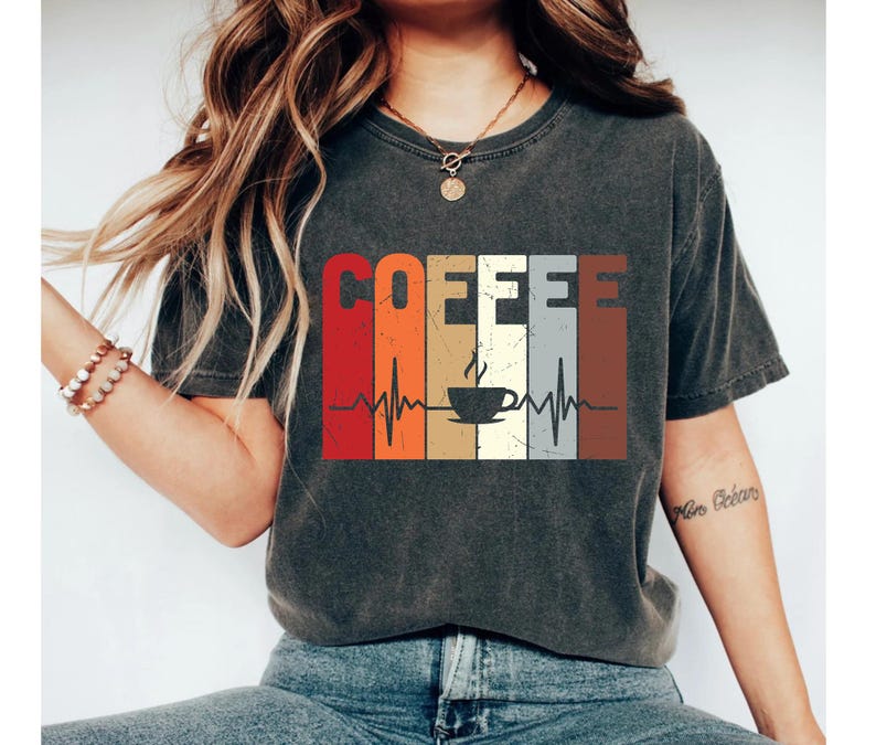 May include: A dark gray t-shirt with a vintage-style graphic of a coffee cup and the word "COFFEE" in a rainbow of colors. The graphic also includes a stylized heartbeat.